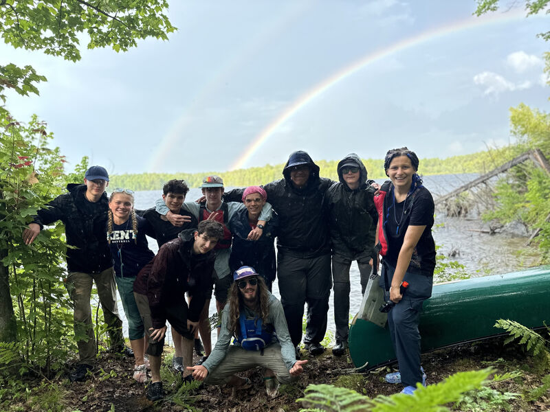 The image shows a group of people posing near a lake with a rainbow in the background. They are dressed in outdoor clothing, suggesting they are on an adventure. A green canoe is also visible. The scene is set in a natural environment, possibly a forest or a park.